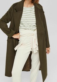 Kobieta w oliwkowo-zielonym długim płaszczu, jasnoszarej koszuli w zielono-białe paski oraz spodni o wysokim stanie w kolorze kremowym, przewiązanych pasującym paskiem.