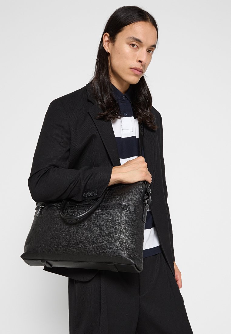 Sac en cuir noir avec une surface texturée, deux poignées et une poche zippée à l'avant, porté sous le bras par un mannequin en costume.