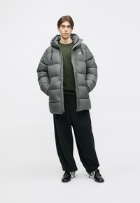 Casaco puffer cinza com capuz, fecho éclair na frente e cordões elásticos, usado por cima de um suéter verde escuro e calças pretas largas.