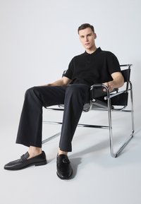 Camisa polo negra y pantalones oscuros usados por un hombre sentado en una silla con estructura de cromo. Los zapatos son negros con un detalle decorativo de hebilla.