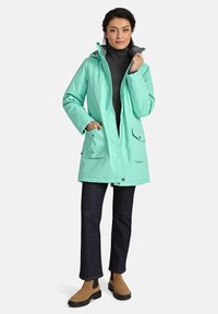 Šviesiai žalias vandeniui atsparus stilingas švarkas su kapišonu, priekyje yra kišenės ir kontrastingas pilkas pamušalas, derinamas su tamsiomis džinsomis ir rudais aulinukais.
