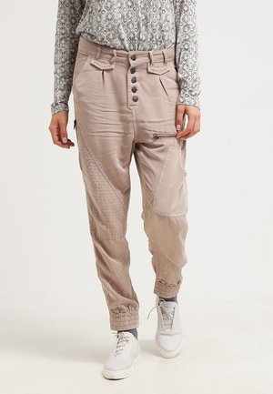 Beżowe spodnie typu cargo z wieloma kieszeniami, zapinane na guziki z elastycznymi mankietami, noszone z wzorzystą, długą bluzką i białymi trampkami.