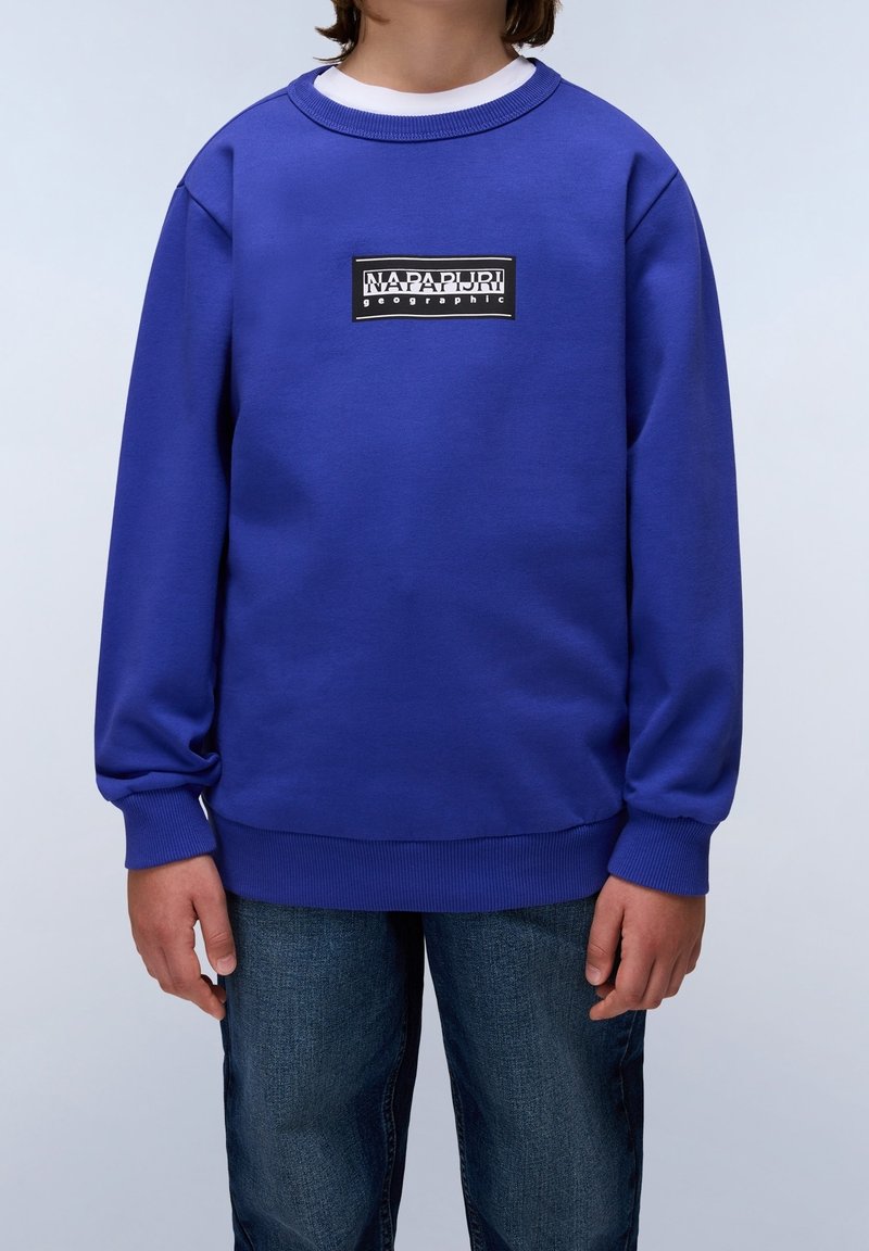 Enfant portant un sweat-shirt bleu Napapijri avec un logo noir et blanc sur la poitrine et un jean bleu, debout devant un fond clair.