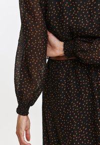 Czarna sukienka z długimi, bufkowymi rękawami, z teksturowanej tkaniny i wzorem małych, nieregularnych kropek w kolorze rdzy. Marszczone mankiety.