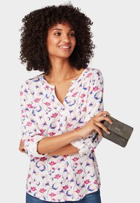 White blouse with pink and blue floral pattern, v-neckline, rolled sleeves, paired with a gray wallet held in hand.