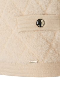 Beiger Pullover mit strukturiertem, flauschigem Oberteil, gerippten Strickbündchen und einer dekorativen schwarzen Knopfdekoration an der Tasche. Goldenes Logo-Tag sichtbar.