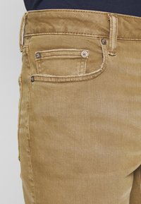 Tan denimjeans med en texturerad yta, med en liten framficka och metallknapppdetalj. Klassisk 5-fickdesign.