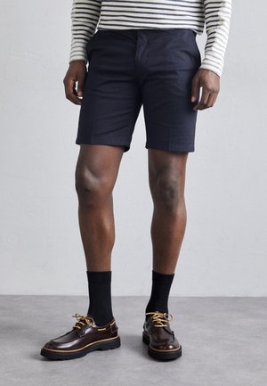 Pantaloni scurți din bumbac bleumarin cu o croială dreaptă, asortați cu pantofi din piele maro și șosete negre. Se vede un tricou cu dungi cu mânecă lungă deasupra pantalonilor scurți.