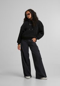 Felpa nera in pile con tasca frontale e logo bianco, abbinata a pantaloni cargo neri a gamba larga con tasche laterali e orli sfilacciati.