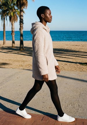 Person går på en strandpromenade iført en beige hættejakke, sorte strømpebukser og hvide sneakers med palmer i baggrunden.