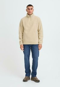 Beige fleece pullover med halv-zip krave, struktureret stof og logo detalje på brystet. Kombineret med blå jeans og brune vandrestøvler.
