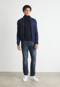 Marinblå zip-up kofta över en marinblå polotröja, ihop med mörka jeans och vita sneakers. Koftan har en subtil logotyp på bröstet.