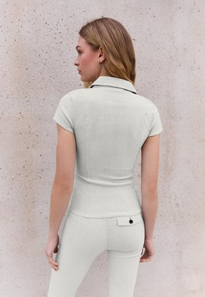 Femeie văzută din spate, purtând un top alb cu mâneci scurte, cu guler și textură rib, și pantaloni asortați, cu buzunar în partea din spate, în fața unui perete texturat deschis la culoare.