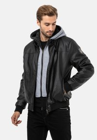 Schwarze Lederbomberjacke mit gerippten Bündchen und Saum, darunter ein grauer Kapuzenpullover und ein dekorativer Aufnäher auf dem linken Ärmel.