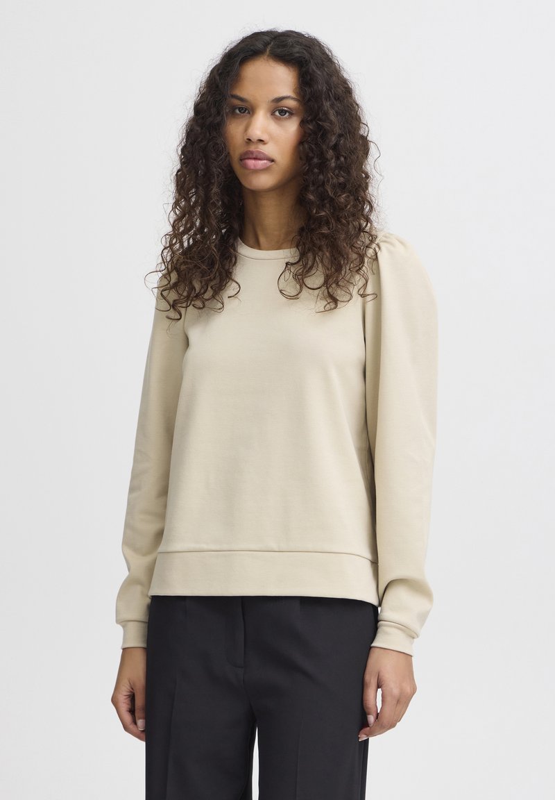 Beige Sweatshirt mit langen Ärmeln und rundem Ausschnitt, ausgestattet mit gepufften Schulterdetails und einem lockeren Schnitt, kombiniert mit dunklen Hosen.