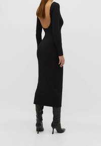 Schwarzes langärmliges Kleid mit tiefem Rückenausschnitt und Spitzenbesatz. Figurbetonte Silhouette mit geradem Saum. Kombiniert mit kniehohen Stiletto-Stiefeln.