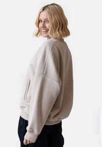 Beżowy, oversize sweter z okrągłym dekoltem, opuszczonymi ramionami i boczną kieszenią. Gładka faktura i minimalistyczny design. Połączony z ciemnymi spodniami.