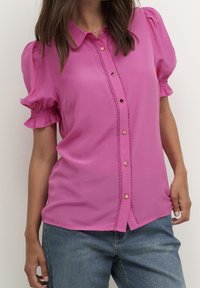 Camisa rosa de botones con mangas abullonadas, con costuras decorativas a lo largo del tapete y botones en tono dorado. Textura de tela suave.