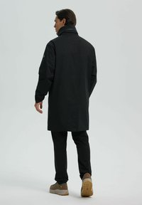 Abrigo negro impermeable con cuello alto y mangas largas, llevado sobre pantalones negros y zapatos marrones. Textura suave, diseño minimalista.