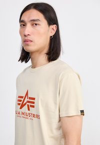 Beige cotton t-shirt featuring a prominent red logo with geometric accents. Short sleeves and a tag on the left sleeve.