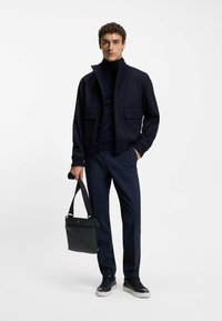 Giacca blu navy con colletto alto e tasche frontali, abbinata a un dolcevita blu navy aderente e pantaloni blu navy su misura; tiene una borsa nera.