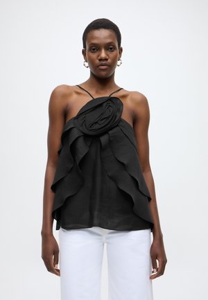 Črna zgornja majica brez rokavov z nagubanim materialom, cvetličnim okrasom na ramenu in lahkotno teksturo, kombinirana z belimi hlačami.