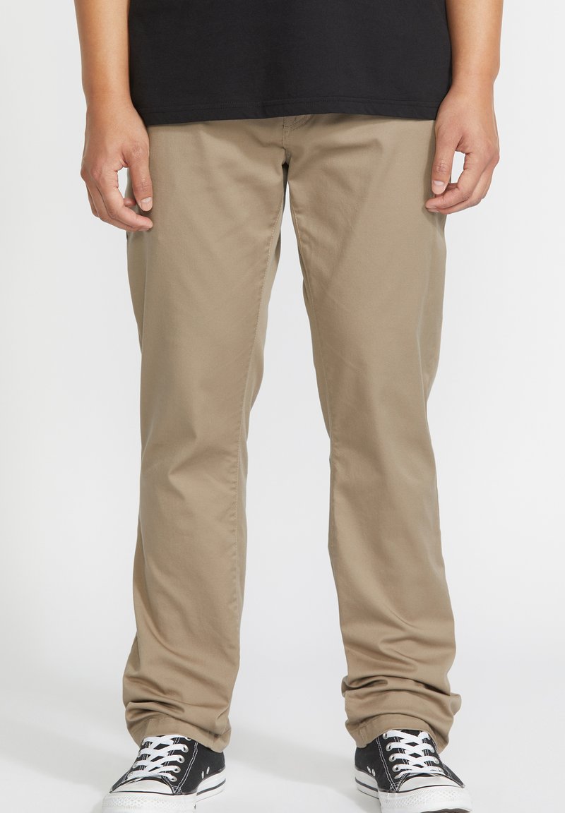 Pantalones de algodón beige de corte recto, con una textura suave y puños enrollados, combinados con zapatillas negras y cordones blancos.
