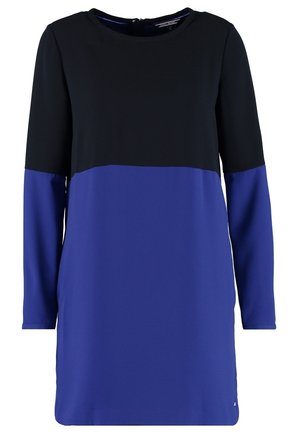 Frau, die ein langärmliges Farbblockkleid in Schwarz und Lila trägt, beige Absätze und eine schwarz-weiße Anhänger-Halskette.