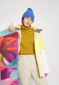 Snowboarder in een geel en witte jack, een mosterdkleurige gebreide trui, beige broeken en een blauwe muts, die een meerkleurige snowboard vasthoudt.