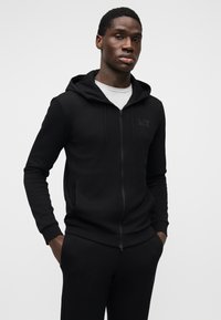 Melna rāvējslēdzēja džemperis ar kapuci no mīksta auduma, ar priekšējo rāvējslēdzēju, divām sānu kabatām un kapuci. Uz krūtīm neliels smalks logotips.