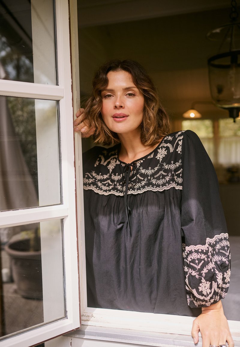 Femme aux cheveux courts et ondulés portant une blouse noire avec une broderie blanche, debout près d'une fenêtre ouverte, regardant calmement droit devant elle à l'intérieur.