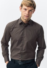 Brown long-sleeve shirt with a geometric pattern, button-down collar, and black buttons. Worn with dark pants and a black belt.