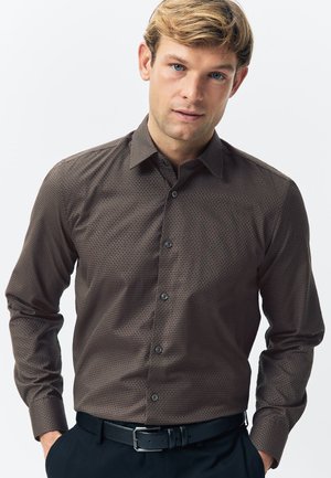 Camisa marrón de manga larga con un patrón geométrico, cuello abotonado y botones negros. Se lleva con pantalones oscuros y un cinturón negro.