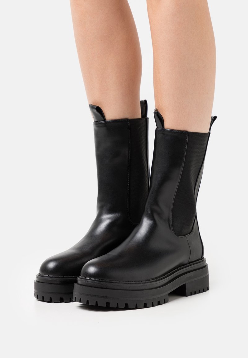 Bottes en cuir noir mi-mollet avec des semelles crantées portées par une personne aux jambes nues sur un fond clair uni.
