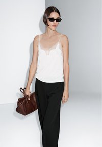 Camisola bianca con pizzo, abbinata a pantaloni neri a gamba larga. Il modello tiene in mano una borsa in pelle marrone e indossa occhiali da sole neri.