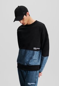 Musta ja sininen väri estetiikkasweatshirt, joka on valmistettu pehmeästä kankaasta, varustettu pyöreällä kaula-aukolla ja brodeeratulla logolla. Yhdistetty samansävyisten farkkujen kanssa.
