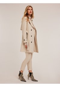 Trench coat bej cu față dublu-zippată, nasturi negri și guler stratificat. Asortat cu pantaloni slim bej și cizme până la gleznă cu imprimeu de șarpe negru și bej.