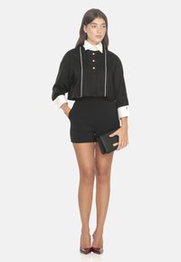 Blusa nera corta con colletto bianco, caratterizzata da bottoni dorati e pieghe, abbinata a pantaloni corti neri a vita alta e una pochette nera.