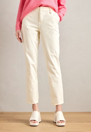 Žena v krémových kalhotách s rovným střihem, bílých otevřených botách na platformě a růžovém svetru stojí na dřevěné podlaze.