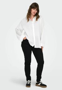 Camicia bianca a maniche lunghe con bottoni, dal fit rilassato, abbinata a pantaloni neri. L'outfit presenta scarpe da ginnastica nere con dettagli bianchi.