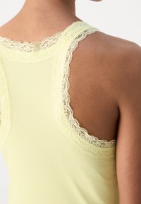 Närbild av en persons rygg som bär en ljusgul ribbad linne med delikat spetskant runt  ärmhålet och halsringningen.