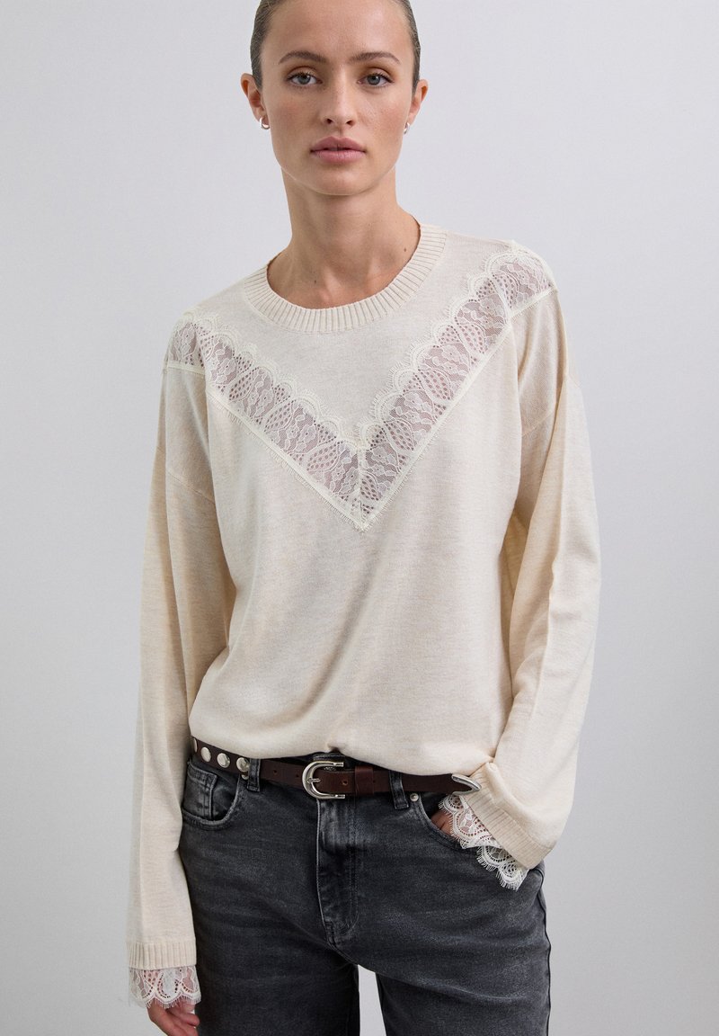 Frau in einem beigen Pullover mit Spitzen-Details an der Brust und den Bündchen, kombiniert mit dunklen Jeans und einem schwarzen Gürtel mit silbernen Nieten.