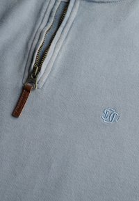 Lichtblauwe pullover met een kwart ritssluiting, van een gestructureerde stof, met een bronzen rits en een klein geborduurd logo in lichtblauw.