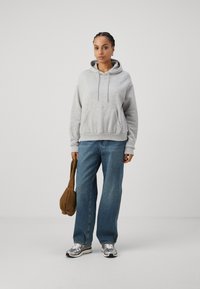Light gray hoodie with a front pocket, drawstring hood, and ribbed hem paired with loose-fit blue jeans and metallic sneakers.