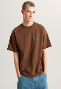 Bruine katoenen t-shirt met korte mouwen, voorzien van een kleine multikleurige afbeelding op de borst, gecombineerd met lichtblauwe denimshorts.