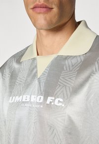 Sølvtrøye med et teksturert mønster, kremfarget krage og hvit "UMBRO F.C." tekst. Inkluderer koordinater på framsiden.