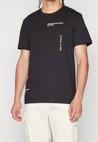 Svart bomullströja med vit textdetalj. Rundhalsad design med korta ärmar och avslappnad passform. Inga mönster eller grafik.