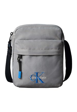 Siva torbica Calvin Klein čez ramo z sprednjim žepom z zadrgo, črnim nastavljivim trakom in modro vezenino z logotipom "cK" spredaj.