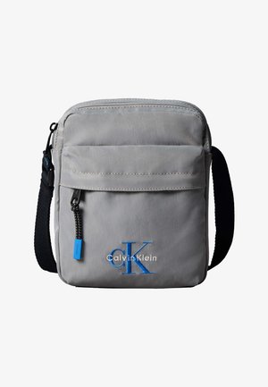 Sivá crossbody taška Calvin Klein s predným vreckom na zips, čiernym nastaviteľným popruhom a modrou výšivkou loga "cK" na prednej strane.