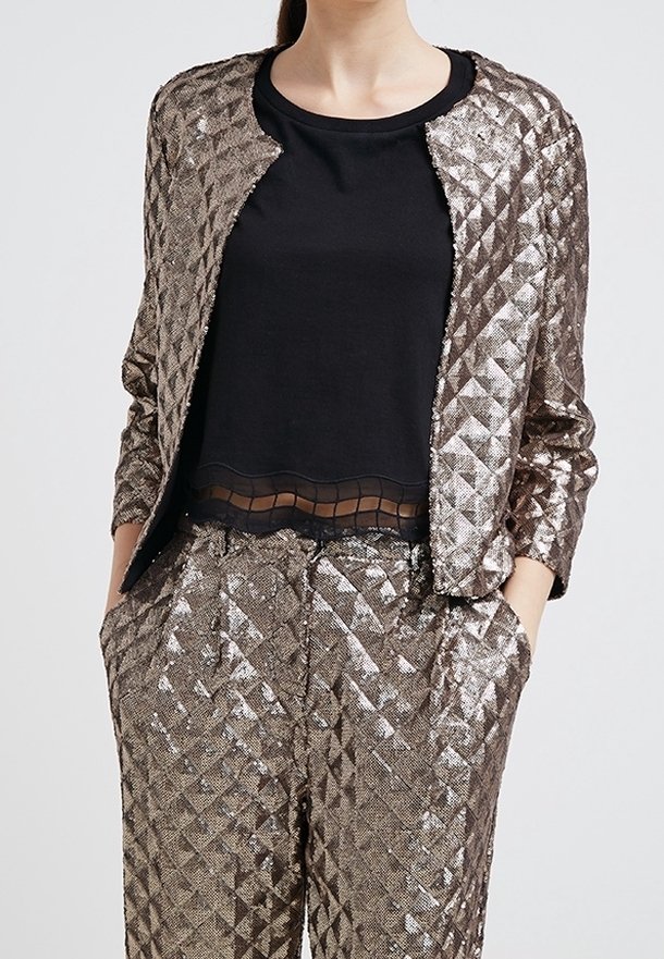 Femme portant une veste à texture métallique et un pantalon assorti, les mains dans les poches, avec un haut noir à ourlet transparent.